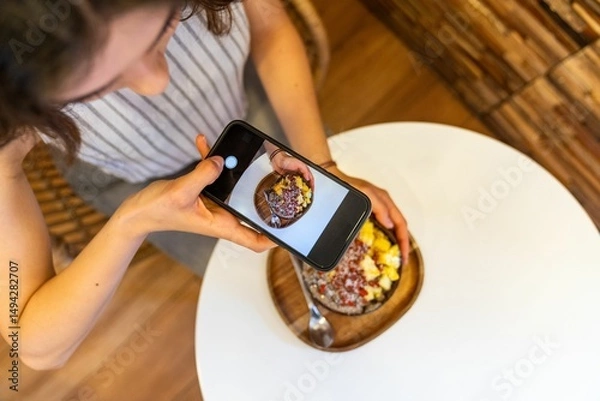 Obraz Woman taking photo of acai bowl with smartphone in cafe.