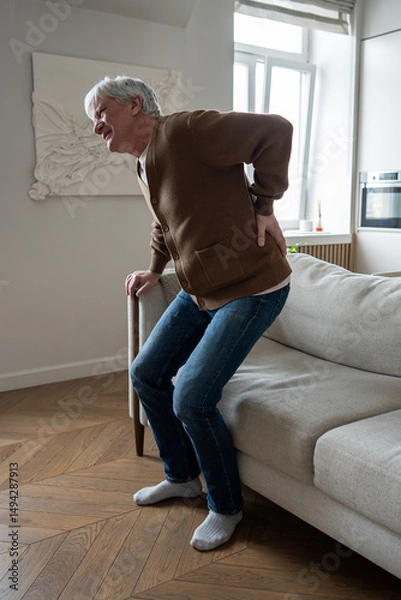 Fototapeta Ill senior man clutches painful spine unable to stand from couch due to multiple health issues, suffering from hernia. Herniated disc agony, pinched nerves, chronic kidney disease, mobility loss. 