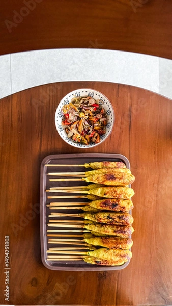 Fototapeta Indonesian satay lilit is served on skewers with traditional sambal matah. It is a delicious Balinese dish presented on a wooden table in a top-down view.