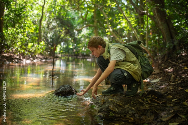 Fototapeta Young boy crouches at the edge forest stream connection between a  learning from the environment holiday ecotourism concept.