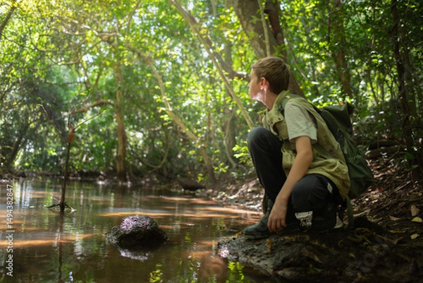 Fototapeta Young boy crouches at the edge forest stream connection between a  learning from the environment holiday ecotourism concept.