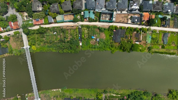 Fototapeta Top-down drone view of a narrow canal bordered by lush greenery and adjacent residential villas with gardens, illustrating Vietnam’s blend of waterways and suburban housing.
