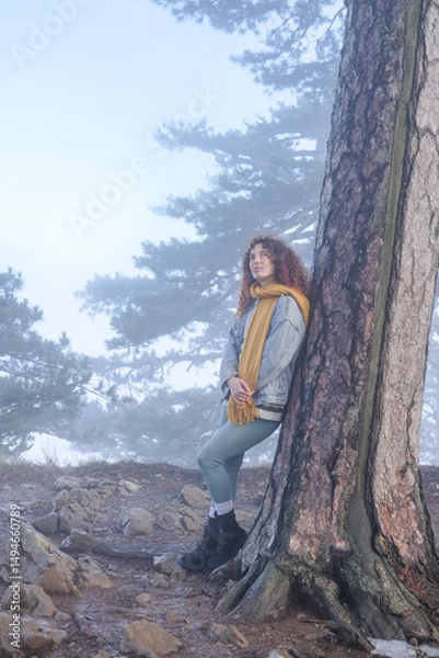 Fototapeta A young female traveler standing in a misty forest, surrounded by tall trees and dense fog. She wears a backpack and outdoor clothing, blending into the mysterious, atmospheric landscape. 