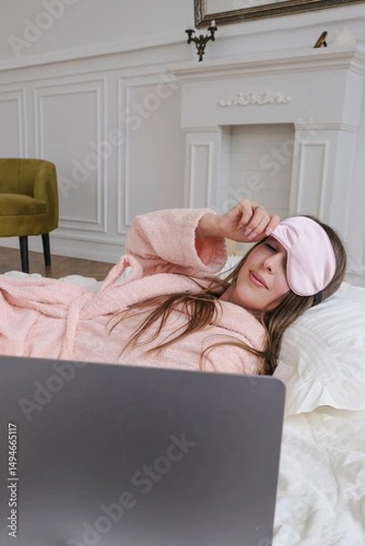 Fototapeta A woman lying in bed and looking at her laptop in the early morning. Wrapped in a cozy blanket with natural light softly filling the room, she starts her day with a calm, relaxed mood. 