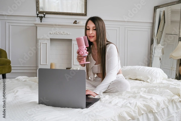 Fototapeta A young woman podcaster records audio content using a professional microphone and laptop while comfortably working from her bed. This intimate home studio setup with soft bedding illustrates modern di