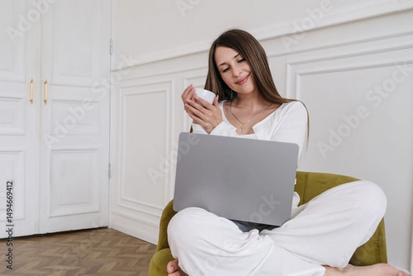 Fototapeta A female freelancer enjoys her coffee while working on a laptop in a comfortable armchair, demonstrating a productive yet relaxed work-from-home setup. Warm natural lighting highlights her modern home