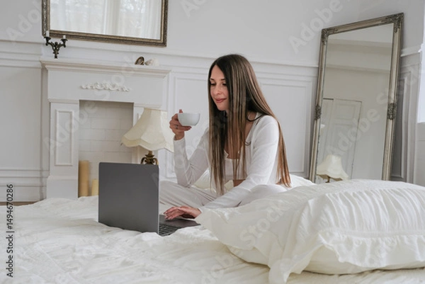 Fototapeta A young female freelancer enjoys her morning coffee while working on a laptop in bed, showcasing a comfortable remote work lifestyle. Sunlight creates a warm atmosphere in this relaxed home office set