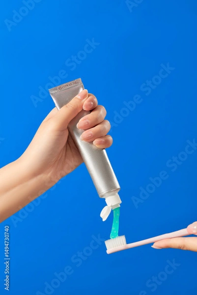 Fototapeta A close-up image of a hand squeezing toothpaste from a metallic tube onto a white toothbrush. The background is bright blue, and the toothpaste is white and clean. 