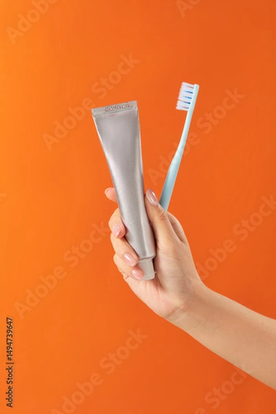 Fototapeta A hand is holding a metallic toothpaste tube and a white toothbrush. The orange background makes the items pop and adds warmth. This image for advertising dental products or personal hygiene visuals.
