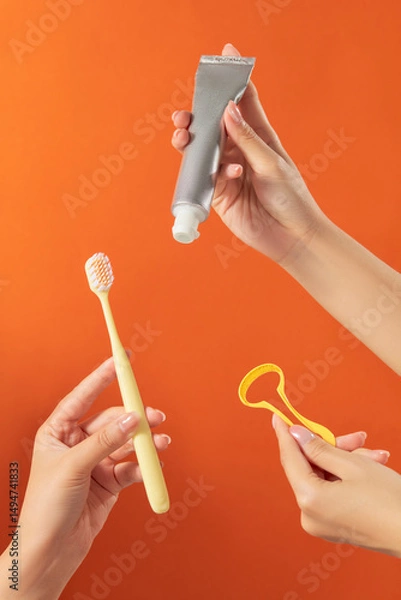 Fototapeta Two hands are holding different oral care tools: a toothpaste a floss pick. The background is orange, giving a vibrant and energetic feel. The photo shows complete daily dental care essentials