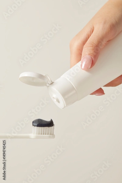 Fototapeta A white hand squeezes a tube of black toothpaste onto a toothbrush. The black color implies a charcoal-based or natural product. Set against a light beige background, the image is clean.