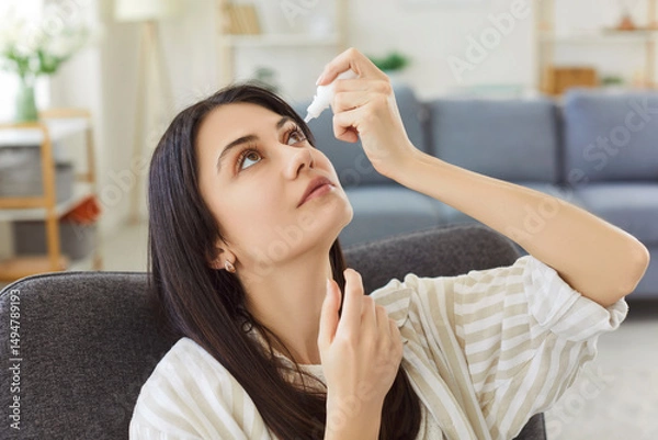 Fototapeta Focused young woman leaning back in armchair, holding dropper and applying eye drops. Female treating dry, irritated or inflamed eyes, focusing on health care routine and maintaining eye moisture.