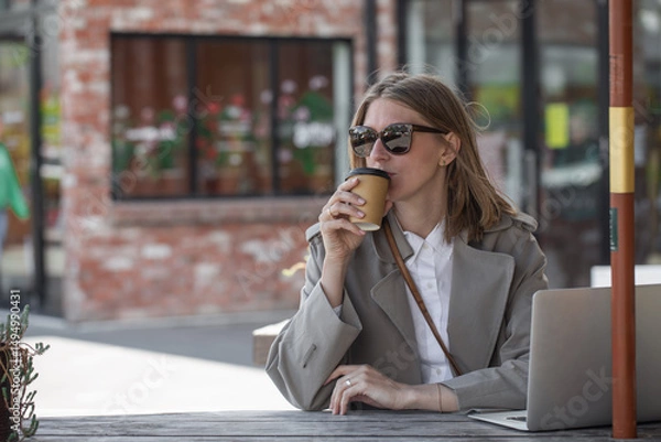 Obraz Caucasian businesswoman drinks coffee and works on laptop, sitting at table outdoor. Remote work