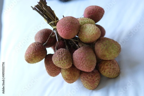 Obraz Close-up of a lychee bunch hanging on a branch, rough red-pink skin and thin stems in humid lighting. . High quality photo