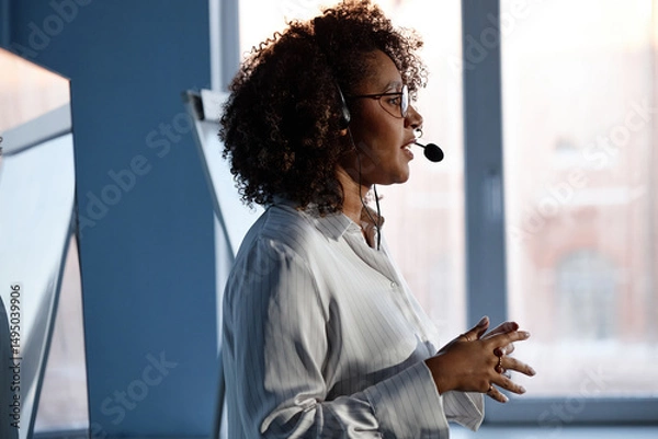 Fototapeta Side view of young African American female expert wearing headset microphone giving presentation at tech conference captured against window in auditorium
