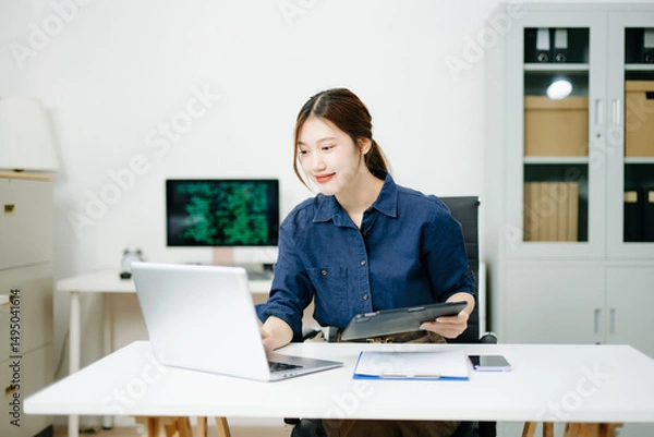 Fototapeta Young female programmer working with tablet and screen displaying code. Modern tech office setup, great for software, coding, IT