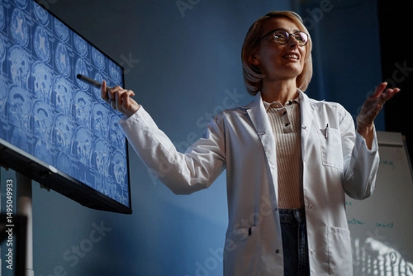 Fototapeta Medium full shot of female head physician presenting lecture on innovative treatment method, pointing at screen displaying brain MRI scan during professional conference in clinic auditorium