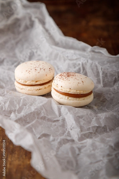 Obraz beige macaroons with chocolate filling, coffee or cappuccino flavor and cocoa powder on paper, wooden background