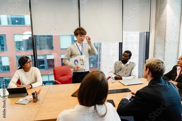 Fototapeta Male intern standing and holding clipboards while talking to a group of five employees sitting at a table in an office