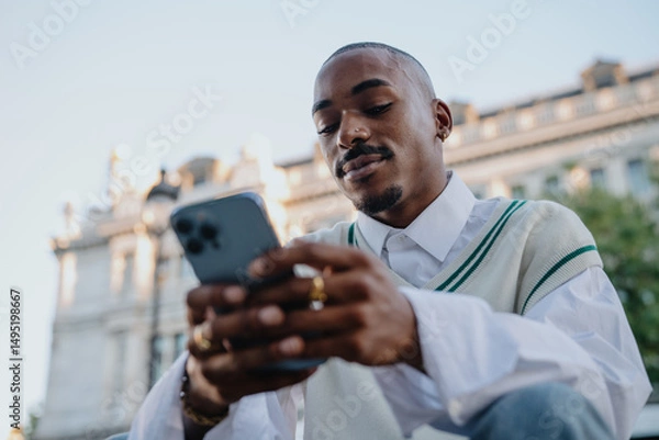 Fototapeta focused Black man in a preppy outfit sits outdoors, scrolling on his smartphone. His stylish look, rings, and well-groomed appearance reflect a modern, sophisticated urban lifestyle