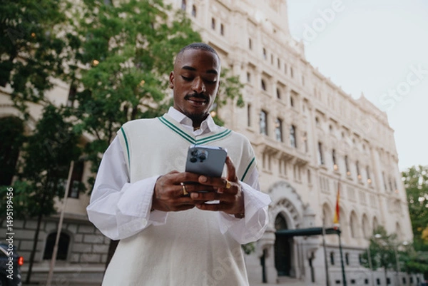 Fototapeta fashionable young African American man uses his smartphone on a city street, focused on his screen. classic architecture behind dark skinned male contrasts with the modern technology in his hands