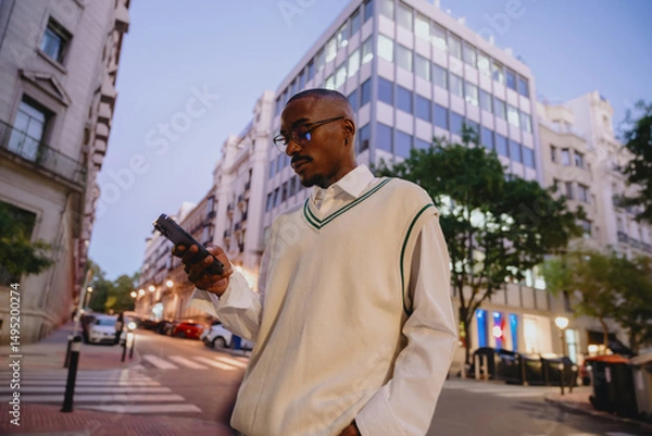Fototapeta young Black man in a stylish white vest and glasses walks through a modern city at night, checking his smartphone. The illuminated buildings and streetlights create a vibrant urban atmosphere