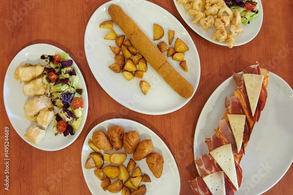 Fototapeta A table with a variety of food items including a plate of chicken wings, a plate of potato wedges, a plate of salad, and a plate of meat