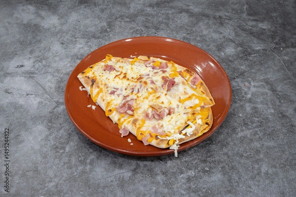 Fototapeta A slice of pizza on a plate. The pizza is topped with cheese and ham. The plate is red and the pizza is on a grey surface