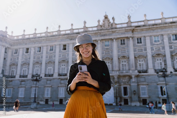 Fototapeta Cheerful young tourist using mobile phone near famous Madrid palace, enjoying solo travel and discovering new places with help of apps, maps, tickets and digital tools on vacation