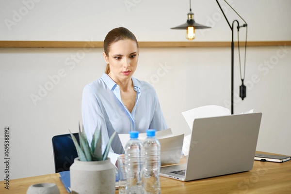 Fototapeta Focused woman reviewing documents at a modern desk setup with a laptop in a well-lit office