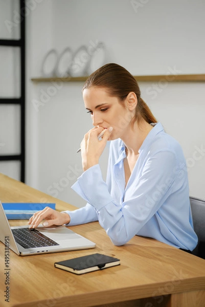 Fototapeta Young woman working intently on a laptop at a wooden desk in a modern office space