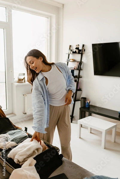 Fototapeta Woman standing and smiling while packing things into a suitcase in the living room