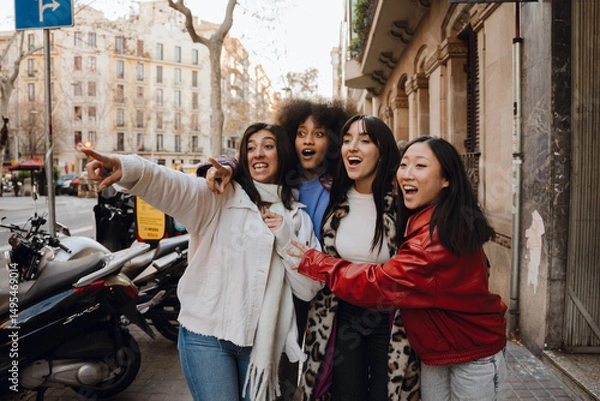 Fototapeta A group of four female friends stand and look in surprise in the direction one of them is pointing, on the street