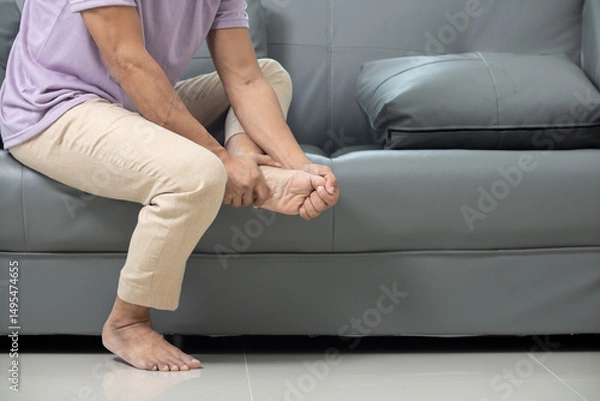 Obraz A young man sits on a sofa and uses his hand to grab his ankle after a home accident, causing ankle pain. A young man has an ankle muscle injury after an accident in his own home.