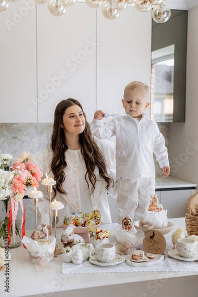 Fototapeta Happy mother and son in white clothes in kitchen with Easter decor. Basket, rabbit, eggs, milk and flowers on table