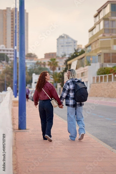 Fototapeta Couple walking hand in hand along seaside promenade at sunset