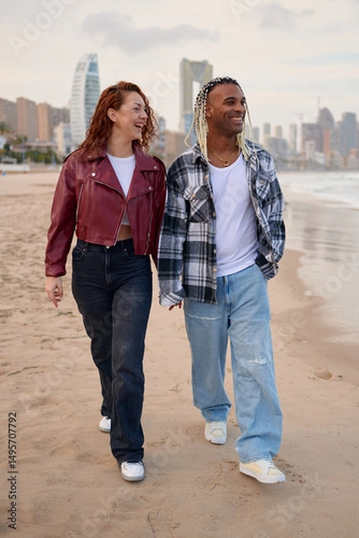 Fototapeta A couple strolls along the sandy beach, smiling and holding hands