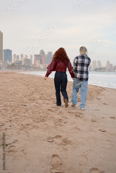 Fototapeta Couple walks hand in hand along sandy beach during sunset near city skyline
