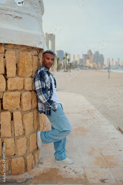 Fototapeta Stylish individual leaning against a stone wall by the beach at sunset