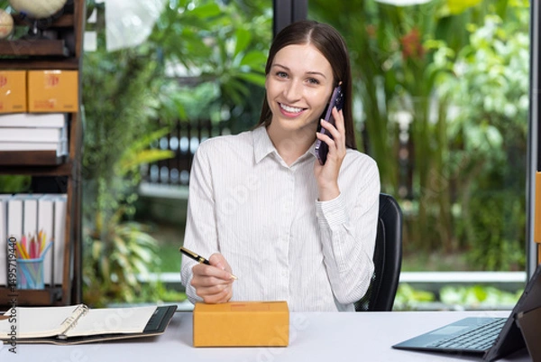 Obraz Smiling young woman talking on phone while preparing a package for delivery at her home office. Concept of online business, e-commerce, and remote work.