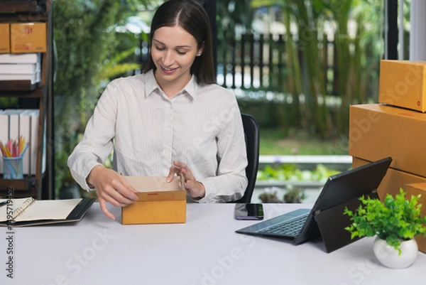 Obraz A young woman packing a cardboard box at her desk in a home office. E-commerce entrepreneur preparing orders for online shipping. Small business startup concept.