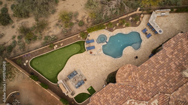 Fototapeta an aerial view of a desert landscaped backyard featuring a travertine tiled pool deck, putting green, outdoor kitchen and fireplace.