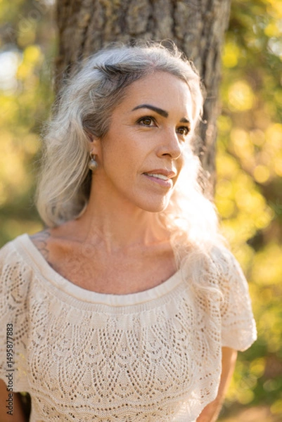 Fototapeta A mature woman with silver hair stands peacefully near a tree, basking in the warm sunlight. Her expression reflects calmness and connection with nature in a serene forest environment.