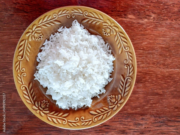 Fototapeta A generous mound of fluffy, white steamed rice is presented on a decorative brown plate with an embossed floral pattern, set against a warm, dark wooden surface. The texture of the rice appears soft