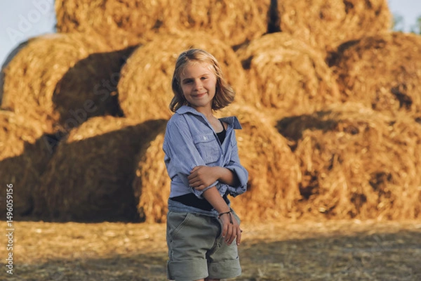 Obraz Horizontal portrait of charming kid girl on the farm posing near rolls of hay bales. Happy childhood in the countryside. Country life, farming and country lifestyle