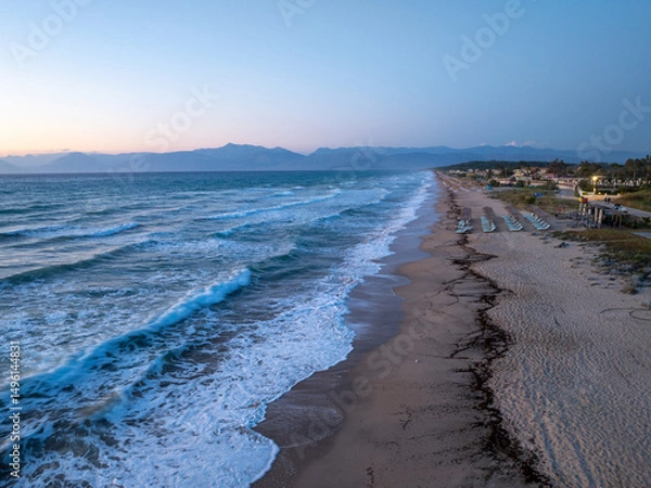 Fototapeta Breathtaking sunset over a tranquil beach with gentle waves washing ashore. The vibrant sky transitions from warm oranges and yellows to deep blues, creating a serene and picturesque coastal landscape