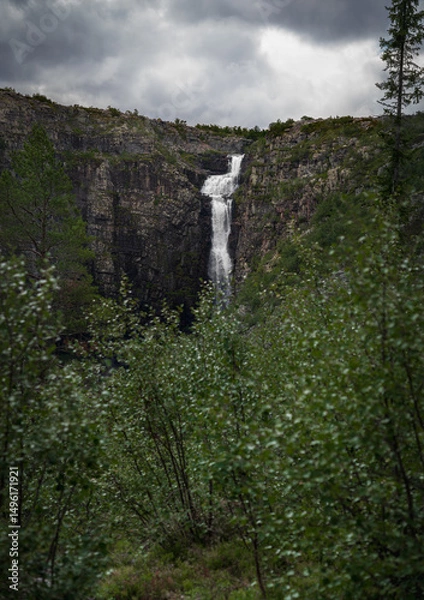 Fototapeta Njupeskär Waterfall flows dramatically down a rugged cliff in Fulufjället National Park, surrounded by dense foliage and under a moody, overcast sky. In Dalarna Sweden
