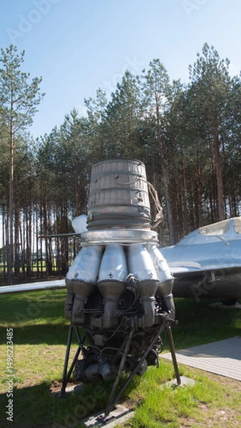 Fototapeta A detailed view of a vintage Soviet rocket engine showcased outdoors near an aircraft, surrounded by lush pine trees under a clear blue sky.