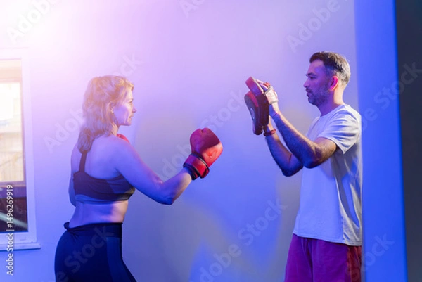 Fototapeta Female boxer training with personal coach using punch mitts in blue-lit gym