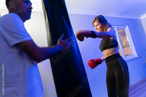 Fototapeta Female boxer training with personal coach using punch mitts in blue-lit gym